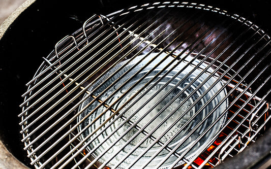 De binnenkant van een Big Green Egg barbecue. Een zilverkleurige aluminium druippan ligt in het midden, geplaatst op een convEGGtor (een keramisch hitteschild). Boven de druippan is een metalen grillrooster bevestigd. De druippan is bedoeld om vet en sappen op te vangen tijdens het barbecueën, terwijl de convEGGtor zorgt voor indirecte hitte. Het geheel is klaar voor gebruik, met gloeiende kooltjes zichtbaar onder de convEGGtor. De setting benadrukt een efficiënte en schone manier van koken.