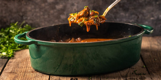 Een donkergroene ovale gietijzeren Dutch Oven van Big Green Egg, gevuld met een rijke stoofpot. Boven de pan wordt een lepel vastgehouden met een portie stoofvlees in een tomatensaus, gegarneerd met verse kruiden. De pan staat op een rustieke houten tafel, en op de achtergrond liggen verse kruiden. De setting straalt een warme, huiselijke sfeer uit, ideaal voor het bereiden en serveren van comfort food.