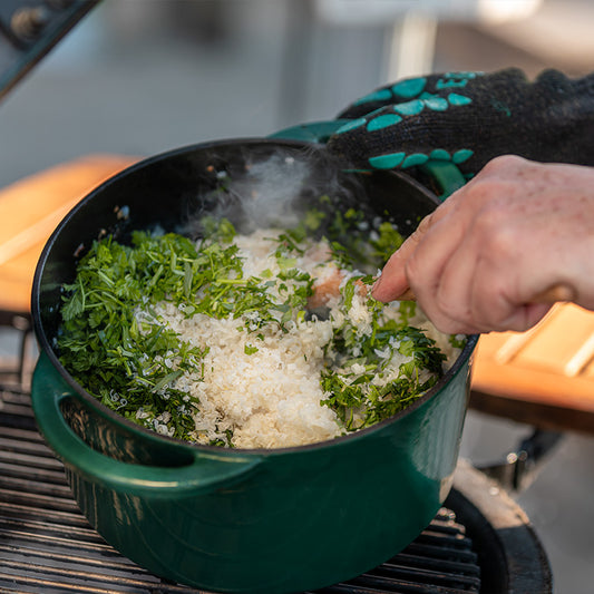Een donkergroene ronde gietijzeren Dutch Oven van Big Green Egg op een Big Green Egg. In de pan wordt een gerecht bereid met verse kruiden, geraspte kaas en rijst of een risotto-achtig mengsel. Stoom komt uit de pan, wat aangeeft dat het gerecht warm is. Het gerecht wordt geroerd door een hand met een houten lepel. De persoon draagt een zwarte hittebestendige handschoen met groene details. Op de achtergrond is een houten werkblad zichtbaar, wat een buitenkeuken-setting suggereert.