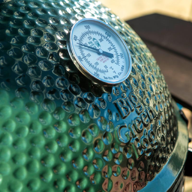 De groene keramische koepel van een Big Green Egg barbecue met een ingebouwde analoge temperatuurmeter. De thermometer is geplaatst in het midden van de koepel en geeft de interne temperatuur van de barbecue aan. De koepel heeft een glanzende, hobbelige textuur met het logo "Big Green Egg" zichtbaar in reliëf. De thermometer is een essentieel hulpmiddel om de temperatuur in de barbecue nauwkeurig te regelen.