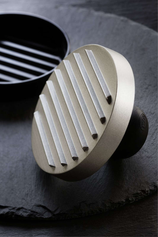 Het onderste, zwarte gedeelte van de hamburgerpers staat op een zwarte, dunne platte steen. Schuin daarvoor ligt op zijn zijkant de RVS pers.