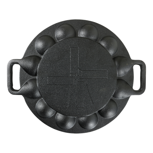 Achterkant van de zwarte, gietijzeren poffertjespan. De pan heeft aan beide zijden een handgreep. Op de vlakke bodem staat het beeldmerk van het merk The Windmill gedrukt. Gefotografeerd recht van boven.