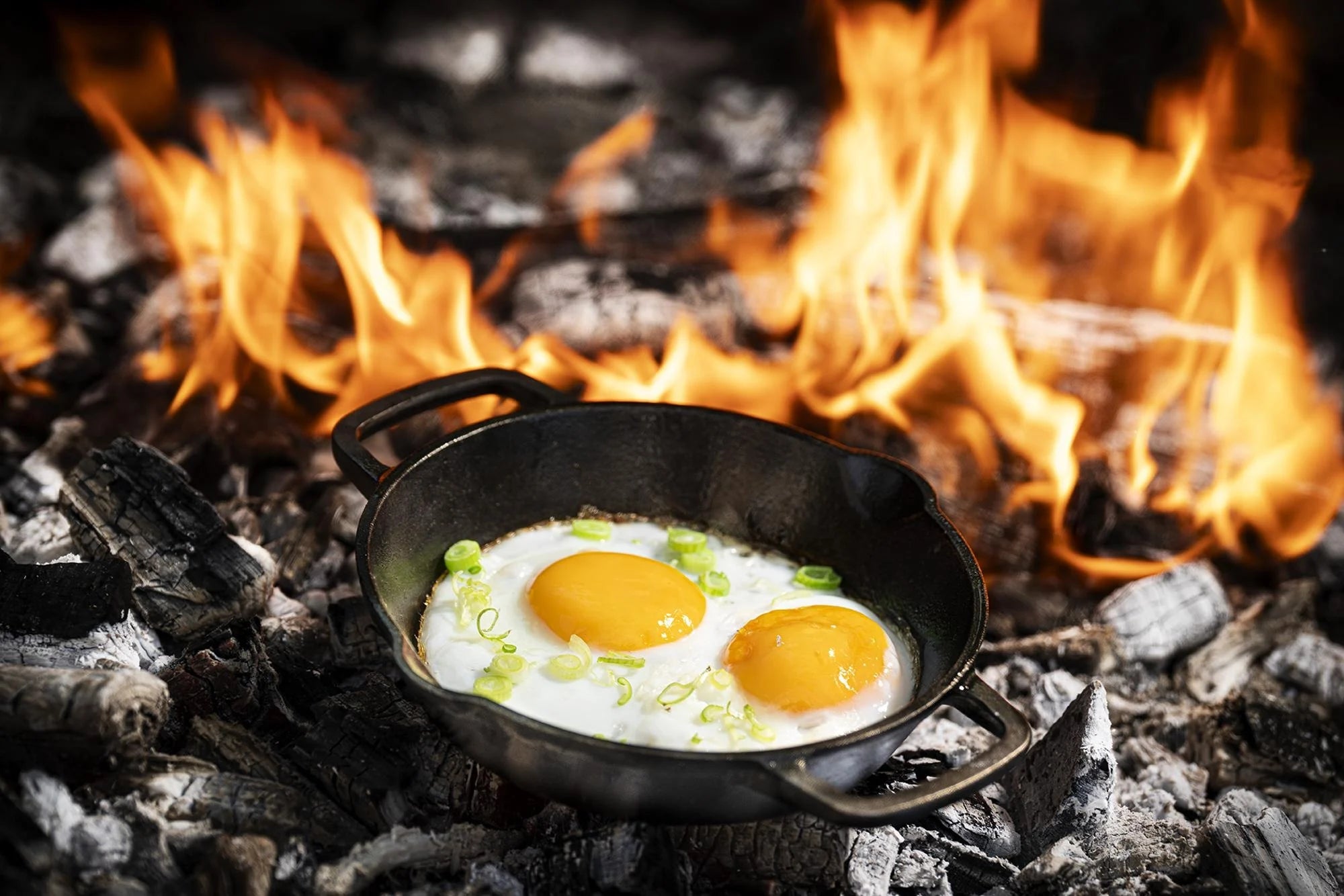 Dezelfde zwarte, gietijzeren pan aan de rand van een open vuur. In de pan ligt een heerlijke uitsmijter te bakken.