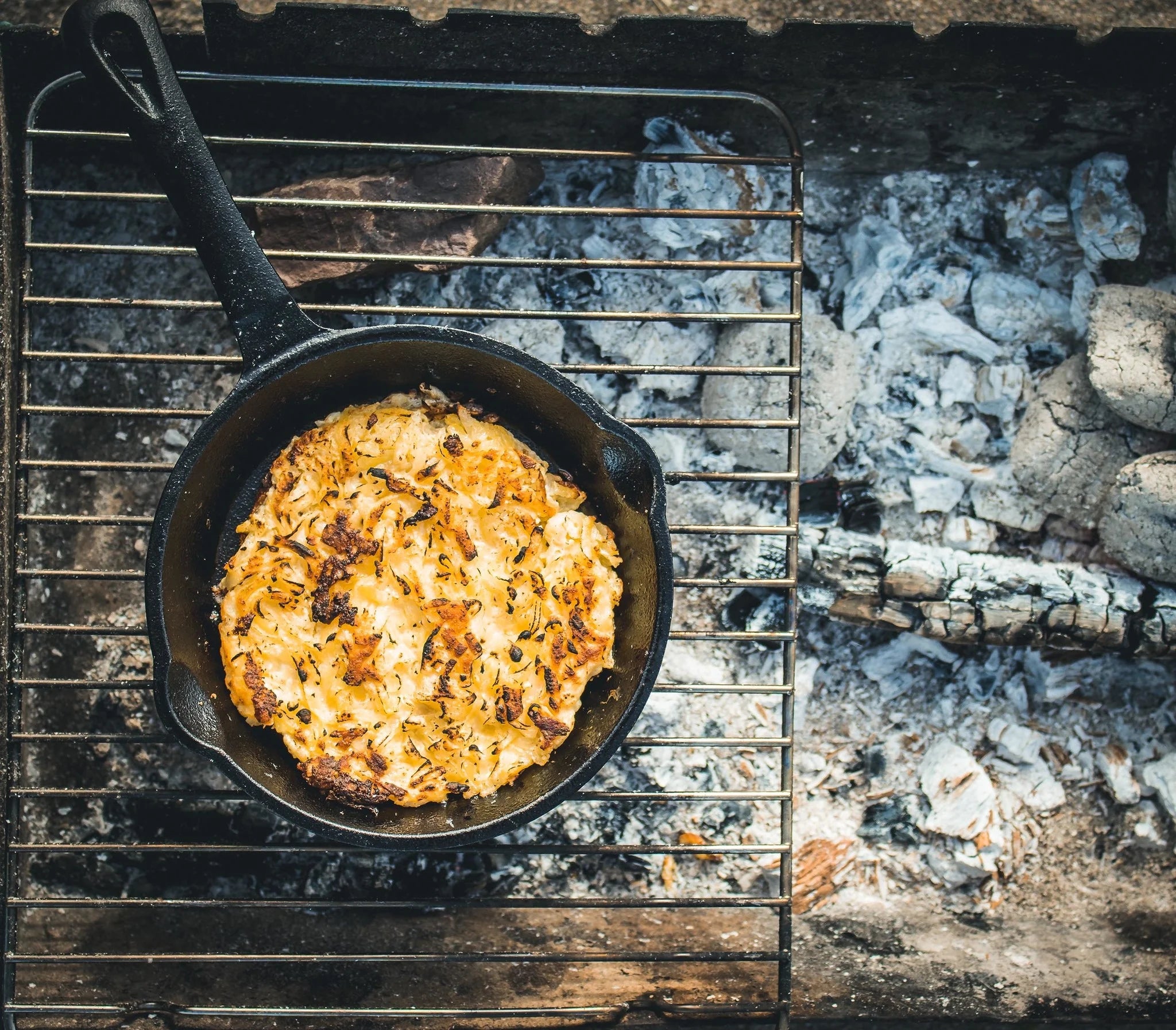 Zwarte gietijzeren skillet met schenktuiten aan beide kanten. De skillet heeft een steel die voorzien is van een ophangoog. De koekenpan is gevuld met een smakelijk uitziende tarte tatin en staat op een grillrooster boven een barbecue.
