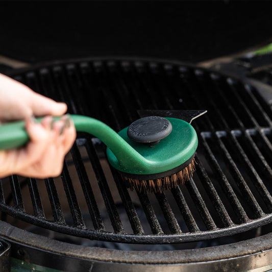 Een schoonmaakborstel van Big Green Egg die wordt gebruikt om een barbecuegrillrooster schoon te maken. Aan de bovenkant van de borstelkop bevindt zich een zwarte knop voor extra druk tijdens het schoonmaken. De borstel wordt vastgehouden door een hand, terwijl het zwarte gietijzeren rooster van de barbecue wordt gereinigd. De setting benadrukt praktisch gebruik van de borstel voor onderhoud van de barbecue.