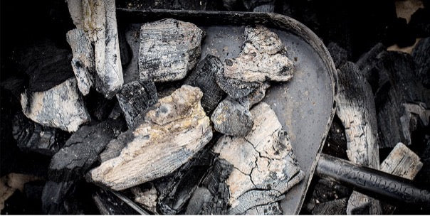 De asschep gelegen in half verbrand houtskool. In de schep ligt ook verbrand houtskool.