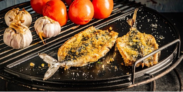 Een Big Green Egg barbecue waarop een halve gietijzeren plancha wordt gebruikt. Op de plancha liggen twee stukken gegrilde vis, bedekt met een krokant paneerlaagje. Aan de zijkant van de plancha zijn gegrilde tomaten en knoflookbollen te zien. De combinatie van de plancha en het rooster maakt het mogelijk om verschillende soorten ingrediënten tegelijk te bereiden. De setting benadrukt veelzijdig koken op de barbecue.