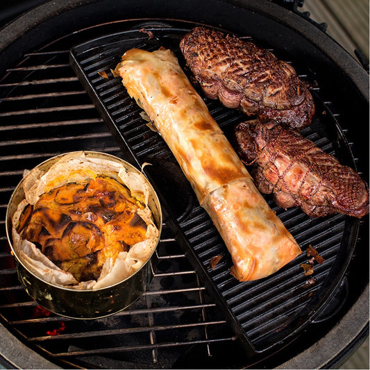 Een Big Green Egg barbecue met verschillende gerechten die worden bereid. Op een halve gietijzeren plancha ligt een mix van ingrediënten: rechtsboven zijn twee stukken gegrild vlees met een mooie korst zichtbaar, in het midden ligt een goudbruin gebakken rol in filodeeg, en linksonder staat een ronde schaal met een gegratineerd gerecht. De plancha heeft een geribbeld oppervlak dat zorgt voor grillstrepen op het vlees. De setting benadrukt veelzijdig koken en bakken op de barbecue.