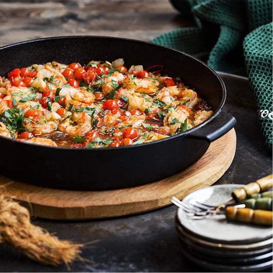 Een gietijzeren pan gevuld met een gerecht bestaande uit garnalen, cherrytomaten, uien, kruiden en een rijke saus. De pan staat op een ronde houten plank, die op een rustieke tafel ligt. Naast de pan liggen stapeltjes kleine borden met vorken, en op de achtergrond is een groene doek te zien. Het gerecht ziet er kleurrijk en smaakvol uit, klaar om geserveerd te worden in een gezellige setting.