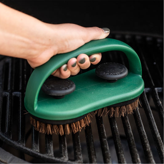 Een hand die een groene handborstel van Big Green Egg gebruikt om een barbecuegrillrooster schoon te maken. De borstel heeft een stevig handvat en twee ronde borstelkoppen De zwarte knoppen bovenop de borstel zorgen ervoor dat de borstelkoppen stevig bevestigd zijn. Het rooster dat wordt gereinigd is van gietijzer gemaakt. De handborstel wordt stevig vastgehouden, wat het praktische gebruik laat zien.