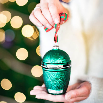 Een vrouw houdt de Big Green Egg kerstbal vast met op de achtergrond een verlichte kerstboom.