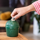 De spaarpot staat op een acacia zijplankje van een Big GreenEgg. Een dameshand stopt een muntje in de sleuf van de spaarpot.