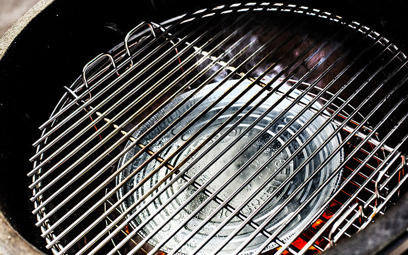 De binnenkant van een Big Green Egg barbecue. Een zilverkleurige aluminium druippan ligt in het midden, geplaatst op een convEGGtor (een keramisch hitteschild). Boven de druippan is een metalen grillrooster bevestigd. De druippan is bedoeld om vet en sappen op te vangen tijdens het barbecueën, terwijl de convEGGtor zorgt voor indirecte hitte. Het geheel is klaar voor gebruik, met gloeiende kooltjes zichtbaar onder de convEGGtor. De setting benadrukt een efficiënte en schone manier van koken.