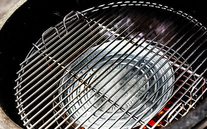 De binnenkant van een Big Green Egg barbecue. Een zilverkleurige aluminium druippan ligt in het midden, geplaatst op een convEGGtor (een keramisch hitteschild). Boven de druippan is een metalen grillrooster bevestigd. De druippan is bedoeld om vet en sappen op te vangen tijdens het barbecueën, terwijl de convEGGtor zorgt voor indirecte hitte. Het geheel is klaar voor gebruik, met gloeiende kooltjes zichtbaar onder de convEGGtor. De setting benadrukt een efficiënte en schone manier van koken.