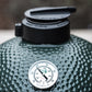 De bovenkant van een groene Big Green Egg barbecue. Op de koepel is een analoge temperatuurmeter gemonteerd, waarmee de interne temperatuur van de barbecue wordt weergegeven. Bovenop de barbecue bevindt zich de rEGGulator, een zwarte gietijzeren ventilatieregelaar. De gegolfde textuur van de koepel en de robuuste accessoires benadrukken de hoogwaardige afwerking van de Big Green Egg. 