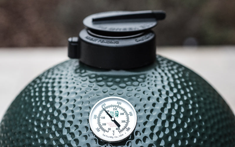 De bovenkant van een groene Big Green Egg barbecue. Op de koepel is een analoge temperatuurmeter gemonteerd, waarmee de interne temperatuur van de barbecue wordt weergegeven. Bovenop de barbecue bevindt zich de rEGGulator, een zwarte gietijzeren ventilatieregelaar. De gegolfde textuur van de koepel en de robuuste accessoires benadrukken de hoogwaardige afwerking van de Big Green Egg. 