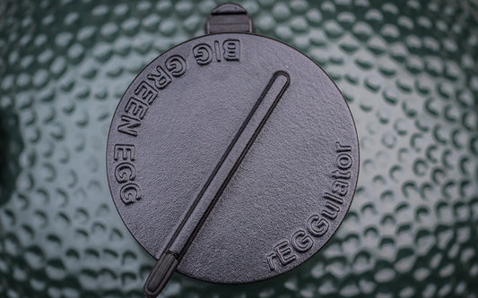 Een close-up van de rEGGulator, een zwarte gietijzeren ventilatieregelaar, gemonteerd op de bovenkant van een Big Green Egg barbecue. Het deksel van de rEGGulator is rond en heeft een geïntegreerde hendel waarmee de luchtstroom kan worden aangepast. Op het deksel staan de woorden "Big Green Egg" en "rEGGulator" in reliëf. De achtergrond laat de karakteristieke gegolfde textuur van de groene keramische koepel van de barbecue zien. 