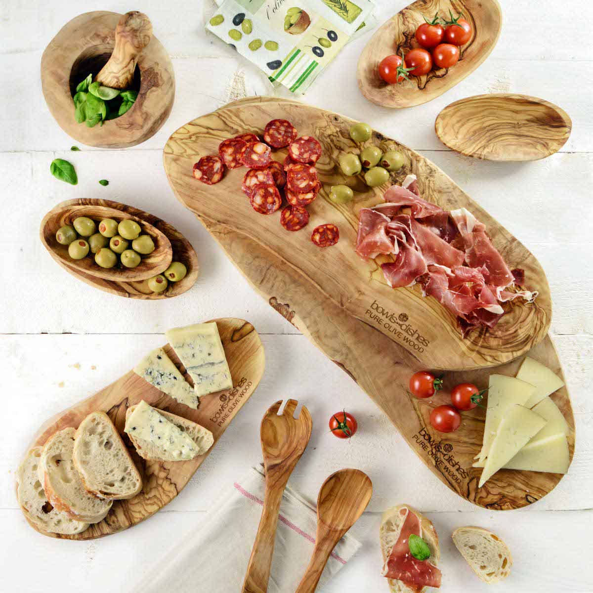 De olijfhouten borrelplank vol met heerlijke borrelhapjes. Om de plank heen zijn ter decoratie andere olijfhouten schaaltjes en planken gezet met lekkere hapjes.
