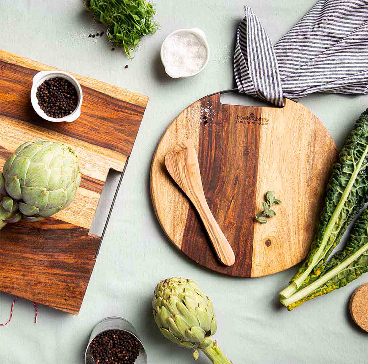 De ronde, houten serveerplank van bovenaf gezien, liggend op een mintkleurige achtergrond. Op de serveerplank ligt een houten lepel met rondom verschillende groentes, schaaltjes met kruiden en een soortgelijke vierkanten houten serveerplank. 