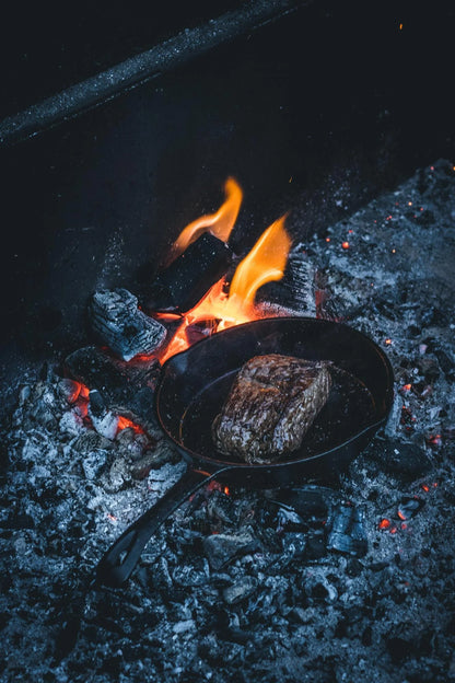 Zwarte gietijzeren skillet met schenktuiten aan beide kanten. De skillet heeft een steel die voorzien is van een ophangoog. De koekenpan is gevuld met een smakelijk uitziende tarte tatin en staat op een grillrooster boven een barbecue.