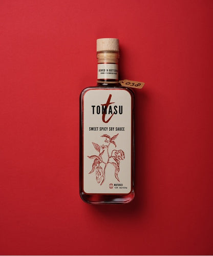 Glazen flesje met Tomasu Sweet Spicy Soy Sauce. Het flesje heeft een dop van kurk en heeft een inhoud van 100ml. Gefotografeerd tegen een ruby rode achtergrond.