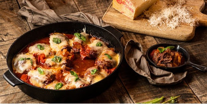 Een gietijzeren pan gevuld met een ovenschotel van gestoofd vlees in tomatensaus, bedekt met gesmolten kaas en verse basilicumblaadjes. Naast de pan staat een kleinere pan met een portie stoofgerecht, gepresenteerd op een servet. Op de achtergrond ligt een houten plank met een blok Parmezaanse kaas en vers geraspte kaas, wat bij de bereiding is gebruikt. De setting is rustiek, met een houten tafel en een linnen doek, wat een warme en huiselijke sfeer creëert.