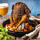 Een hele gegrilde kip met een gekruide korst, rechtopstaand in een gietijzeren pan. De kip is geplaatst op een bierblikhouder, waarbij het blik bier zichtbaar is. Rondom de kip liggen gegrilde groenten. Op de achtergrond staat een glas bier met een schuimkraag op een houten tafel. Het geheel straalt een rustieke en smakelijke barbecue-maaltijd uit, bereid op een Big Green Egg. Er liggen ook een linnen servet en verse kruiden in de voorgrond.