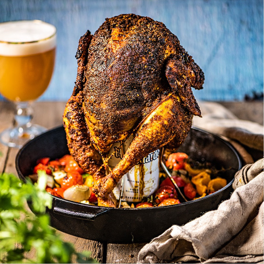Een hele gegrilde kip met een gekruide korst, rechtopstaand in een gietijzeren pan. De kip is geplaatst op een bierblikhouder, waarbij het blik bier zichtbaar is. Rondom de kip liggen gegrilde groenten. Op de achtergrond staat een glas bier met een schuimkraag op een houten tafel. Het geheel straalt een rustieke en smakelijke barbecue-maaltijd uit, bereid op een Big Green Egg. Er liggen ook een linnen servet en verse kruiden in de voorgrond.