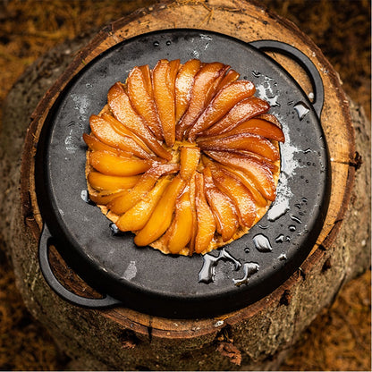 Een ronde, rustieke perentaart in een gietijzeren pan. De taart bestaat uit plakjes gekarameliseerde peren die in een cirkel zijn gerangschikt, glanzend van de karamel. De pan staat op een houten boomstam, wat een natuurlijke en outdoor sfeer geeft. Het gerecht ziet er ambachtelijk en warm uit, ideaal voor een buitenomgeving, bereid op een Big Green Egg.