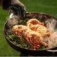 Een gietijzeren pan gevuld met twee grote stukken gegrilde vis, geserveerd op een bed van knapperige groenten. De pan wordt vastgehouden door een persoon die een zwarte barbecuehandschoen draagt, naast een open keramische barbecue van Big Green Egg. Er stijgt stoom op van het gerecht, wat aangeeft dat het net bereid is. De achtergrond is een groene grasveldsetting, wat duidt op een buitensituatie.
