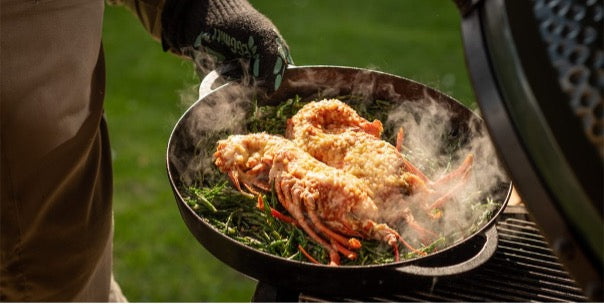 Een gietijzeren pan gevuld met twee grote stukken gegrilde vis, geserveerd op een bed van knapperige groenten. De pan wordt vastgehouden door een persoon die een zwarte barbecuehandschoen draagt, naast een open keramische barbecue van Big Green Egg. Er stijgt stoom op van het gerecht, wat aangeeft dat het net bereid is. De achtergrond is een groene grasveldsetting, wat duidt op een buitensituatie.