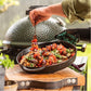 Een persoon die een vleesspies vasthoudt boven een ovale gietijzeren pan met gegrilde groenten, kruiden en vleesspiezen. De pan staat op een houten plank naast een groene keramische barbecue van Big Green Egg. De persoon draagt een zwarte barbecuehandschoen met het woord "EGCMIT". De setting is buiten, waarschijnlijk in een tuin, en straalt een gezellige barbecue-sfeer uit.